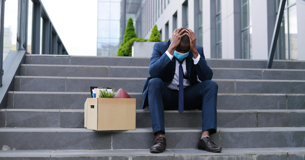 Homem sentado em uma escada com as mãos na cabeça ao lado de uma caixa de pertences em referência ao desemprego.