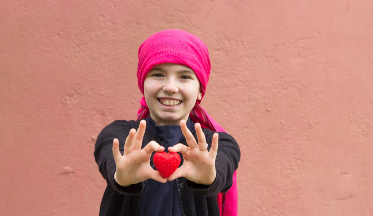 Criança com câncer segurando, com as duas mãos, um pequeno coração de plástico vermelho