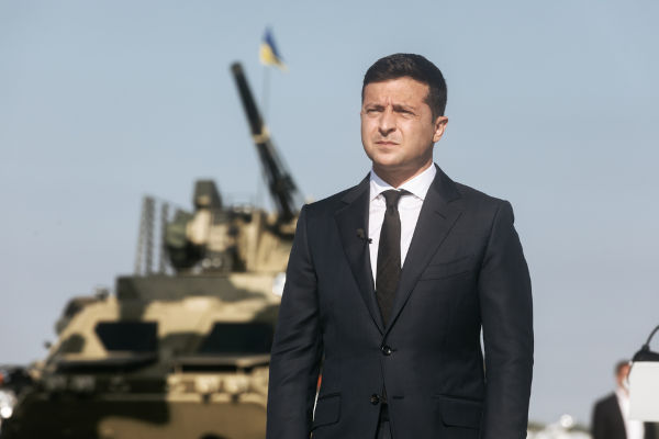 Volodimir Zelensky de terno e gravata com tanque de guerra ao fundo.
