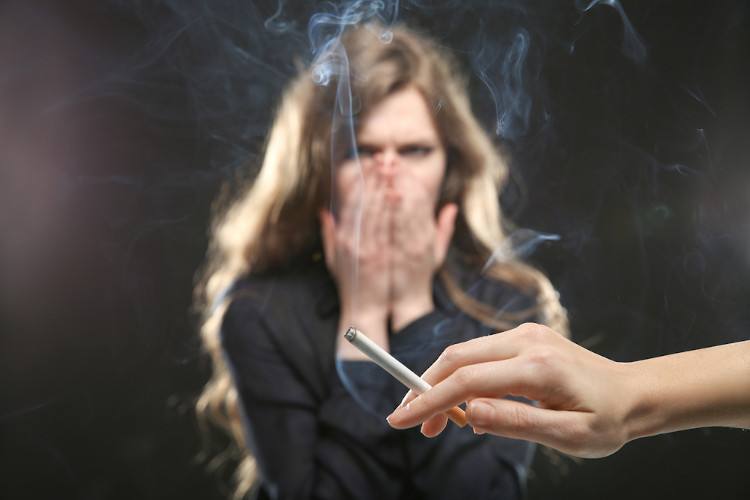 Mão segurando cigarro aceso; ao fundo, mulher tampando o rosto com as mãos e expressando desagrado com a fumaça do cigarro