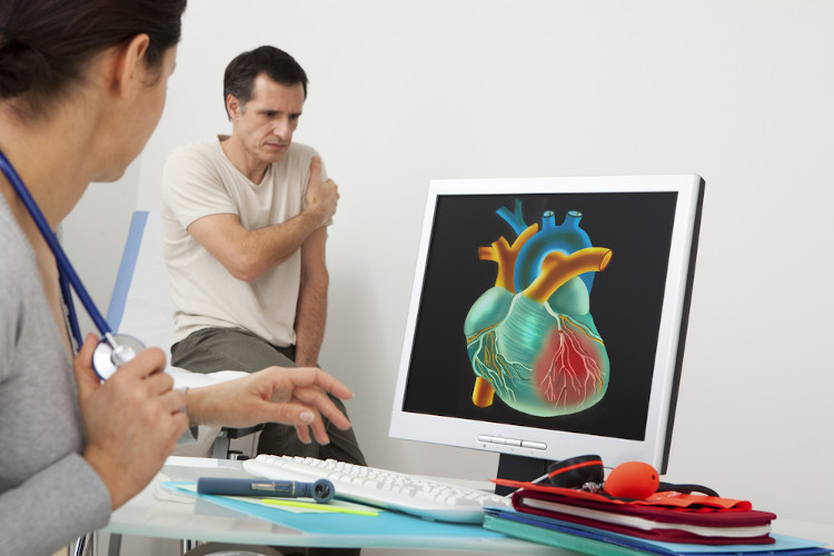 Monitor mostrando imagem de coração humano; médica em primeiro plano, e, ao fundo, homem com mão em braço, indicando dor