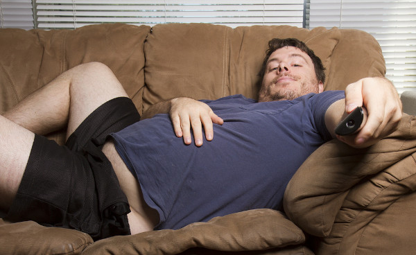 Homem obeso deitado no sofá com controle de televisão na mão