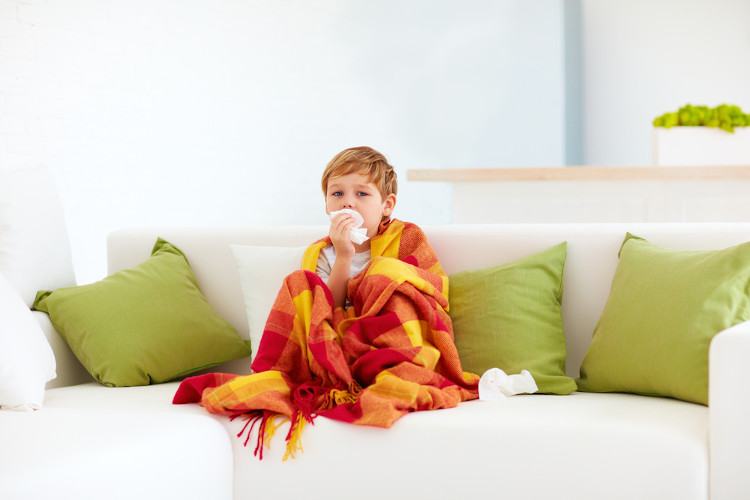 Criança sentada no sofá envolta em uma manta e tampando a boca com papel