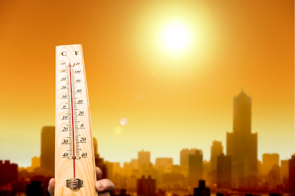 Pessoa segurando um termômetro medindo 50 °C com o Sol ao fundo em uma região urbana.