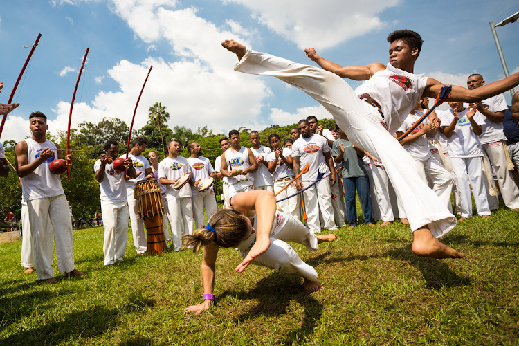 Dois capoeiristas jogando capoeira enquanto os outros capoeiristas batem palma no ritmo do berimbau.