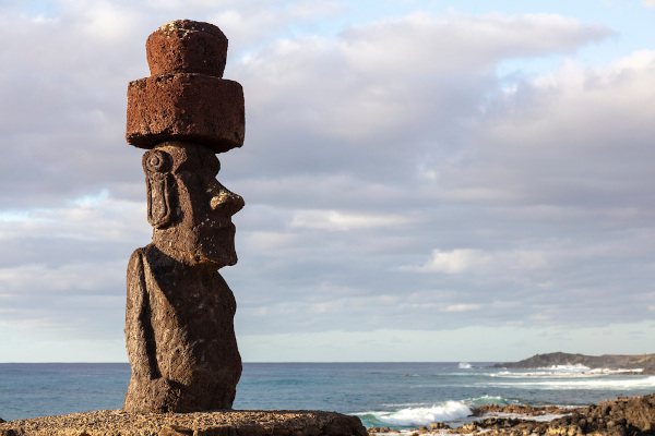Moai na paisagem da Ilha de Páscoa, estátua com forma humana e chapéu feito de uma pedra vulcânica avermelhada.