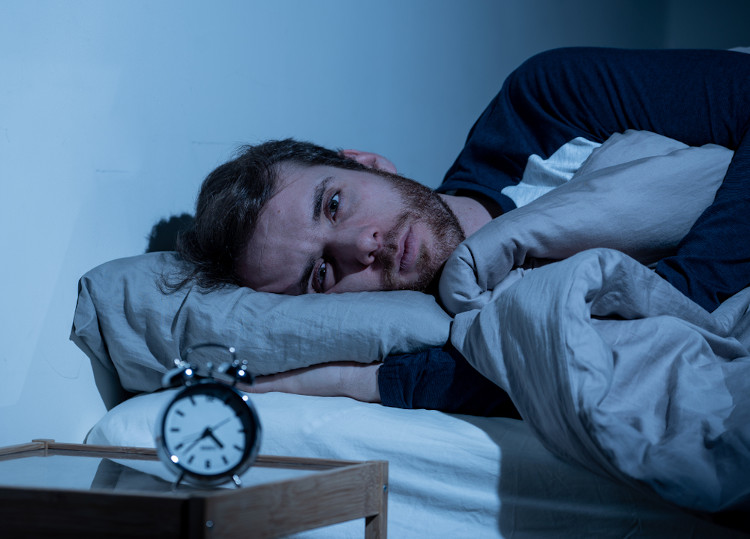  Homem deitado na cama olhando para um despertador que está na mesa ao lado; narcolepsia causa fragmentação do sono.