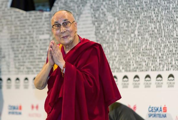 Tenzin Gyatso, o atual Dalai Lama, vestido com roupa vermelha, com as mãos encostadas uma na outra.