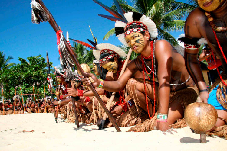 Indígenas brasileiros ajoelhados na areia durante ritual, em texto sobre cultura brasileira.