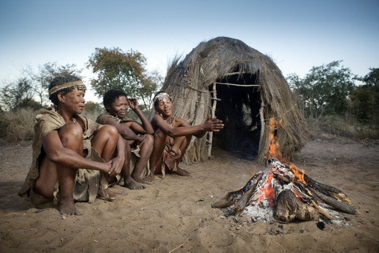 Três pessoas do povo originário africano san sentados próximos a uma fogueira.