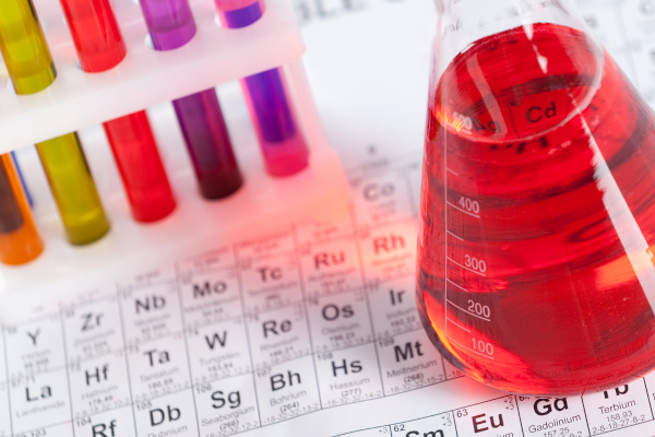 Vidrarias de laboratório contendo líquidos coloridos próximas a uma Tabela Periódica, uma alusão à Química Inorgânica.